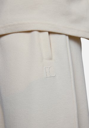 Cream-colored ribbed fabric close-up showing a side pocket with a small embroidered "EC" logo on the garment.
