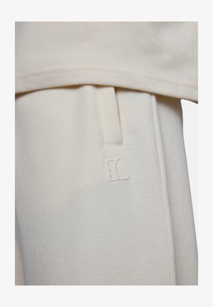 Cream-colored ribbed fabric close-up showing a side pocket with a small embroidered "EC" logo on the garment.