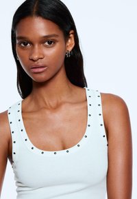 White ribbed tank top featuring rounded neckline with black stud details along the straps. Smooth texture and fitted design.
