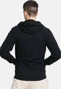 Felpa nera con cappuccio e cordoncino, polsini e orlo a costine. Presenta del nastro decorativo lungo le spalle, tessuto liscio e vestibilità comoda.