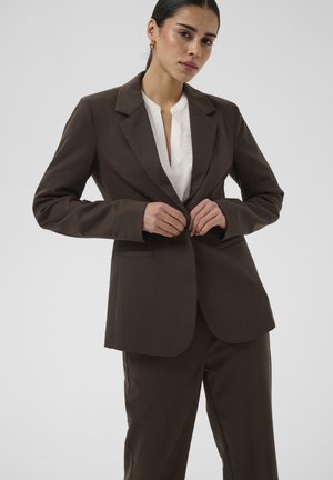Femme boutonnant un blazer marron foncé sur un chemisier blanc, portant un pantalon assorti, sur un fond clair uni.