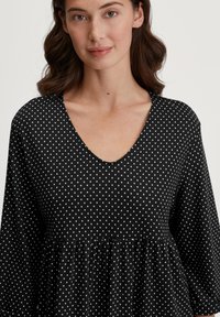 Schwarzes Polka-Dot-Oberteil mit V-Ausschnitt und lockerer Passform, ausgestattet mit einer Raffungsdetails an der Taille. Der Stoff wirkt weich und leicht.