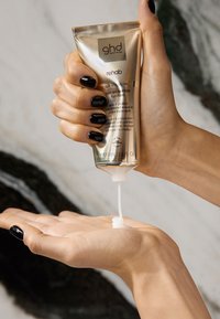 Złota tubka wygładzającego preparatu ghd Rehab Split End Sealer, z którego produkt jest wyciskany na dłoń. Pokazywana jest kremowa, biała substancja o błyszczącej teksturze i gładkiej konsystencji.