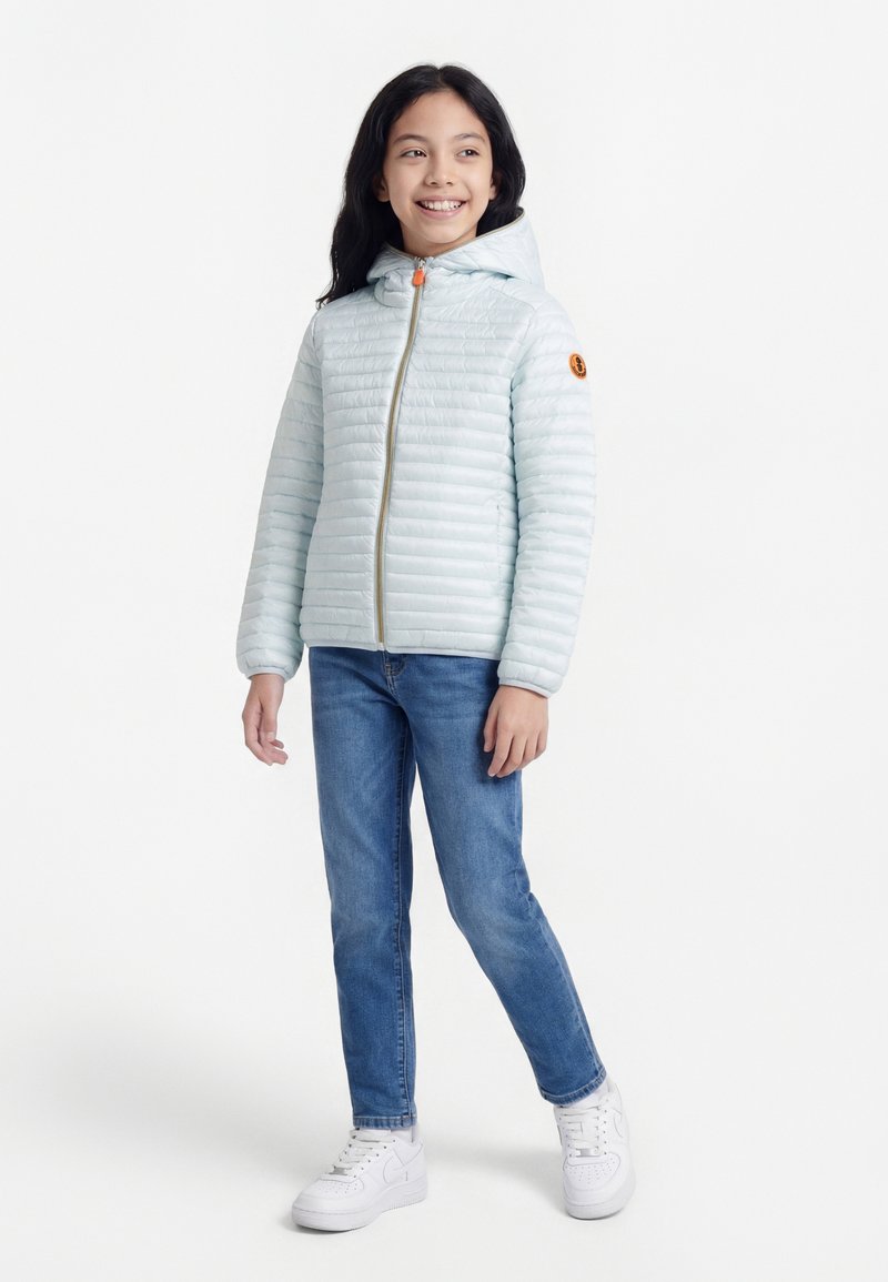 Hosszú sötét hajú lány világoskék steppelt kapucnis dzsekiben, kék farmerben és fehér tornacipőben, állva, mosolyogva, jobbra nézve.