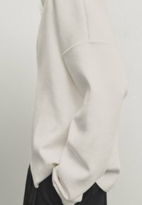 Sweatshirt blanche à manches longues avec une coupe décontractée, texture en tricot, coutures d'épaules tombantes et manches à revers, rentrée dans un pantalon foncé.