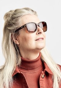 Ruskotona kehyksinen aurinkolasit, joissa on liukuväriset ruskeat linssit. Henkilöllä on pitkät vaaleat hiukset, yllään ruosteenvärinen poolopaita ja takki.