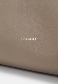Smeđa kožna torba s teksturiranom površinom. Sadrži zlatni logo "Coccinelle", naglašavajući brand. Jednostavan i minimalistički dizajn.