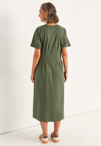 Olivgrön midiklänning med korta ärmar, med veck i ryggen, kombinerad med ljusa sneakers. Slätt bomullsmaterial.
