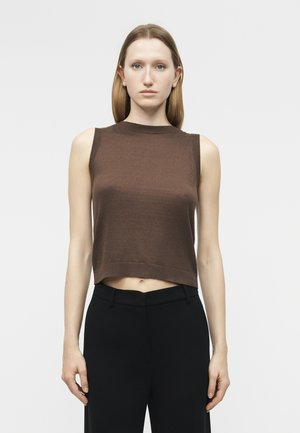 Femme aux cheveux châtain clair et raides, portant un top marron sans manches et court, ainsi qu'un pantalon noir taille haute, debout devant un fond blanc.