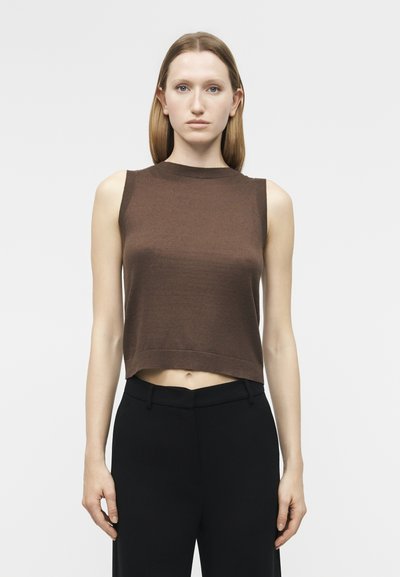 Woman with straight light brown hair wearing a sleeveless brown cropped top and high-waisted black pants standing against a white background.