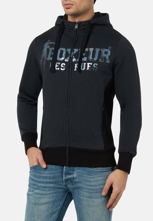 Schwarze Zip-Up-Hoodie mit einem strukturierten, metallischen Logo auf der Brust. Verfügt über verstellbare Kordeln und gerippte Bündchen. Gezeigt mit hellblauen Jeans.