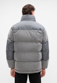 Chaqueta acolchada gris con un cuello alto, que presenta una mezcla de tejidos texturizados y lisos, en diferentes tonos de gris, con un corte relajado.