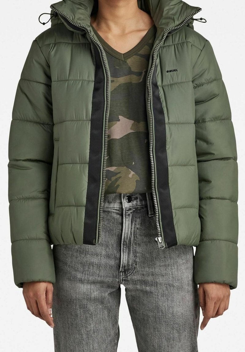 Zielona kurtka puchowa z kapturem, widocznym zamkiem błyskawicznym i czarnymi bocznymi akcentami. Noszona na koszulę w kamuflażu i szare dżinsy. Teksturowany, ciepły design.