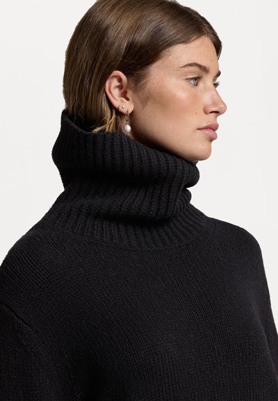 Sort strikket turtleneck sweater med ribbelsstruktur, høj krave og blødt stof. Modellen bærer øreringe og viser en sideprofil.