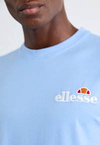 Light blue t-shirt made of soft fabric. Features embroidered "ellesse" logo in white with a red and orange semicircle accent. Short sleeves and crew neck.