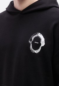 Μαύρο φούτερ με κουκούλα, που διαθέτει λευκό σχέδιο με στόμα καρχαρία και τη λέξη "HUGO" στο κέντρο. Κατασκευασμένο από βαμβακερό υλικό, με άνετη εφαρμογή.