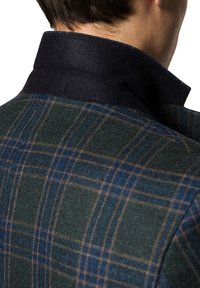 Szkocka wełniana kurtka z ciemnoniebieskim kołnierzem oraz mieszanką zielonych, granatowych i brązowych wzorów w kratę. Widoczna faktura materiału.