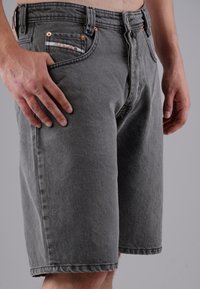 Man wearing gray denim shorts with copper rivets and front pockets, hands relaxed at sides against a plain gray background.