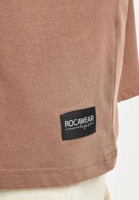 T-shirt in cotone marrone con etichetta nera ROCAWEAR ricamata sull'orlo. Il tessuto ha una consistenza liscia con dettagli a costine.