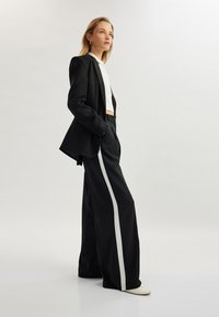Zwarte pak met een op maat gemaakte blazer en wijde broek met een witte zijstreep. De outfit wordt aangevuld met een cropped wit topje en neutrale schoenen.