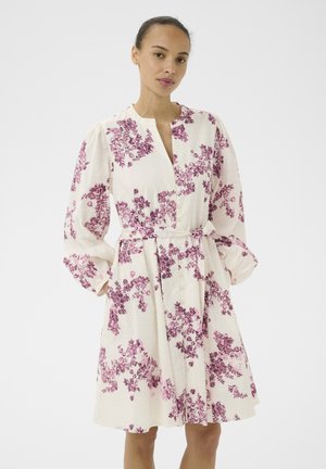 Ženska v belem obleki do kolen z dolgimi nabranimi rokavi in vijoličnim cvetličnim vzorcem, stoji pred enobarvnim ozadjem.