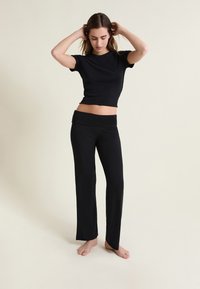 Femme en haut court à manches courtes noir et pantalon noir, debout pieds nus devant un fond clair uni.