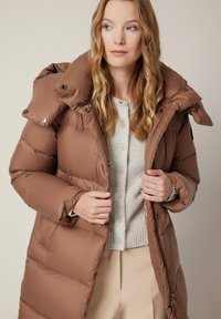 Brązowa puchowa kurtka z wysokim kołnierzem, zapinana na zamek i zatrzaski. Posiada pikowaną fakturę i kieszenie po bokach. Nosi się ją na lekkim swetrze.