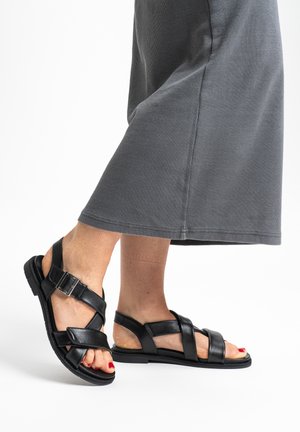 Füße in schwarzen, offenen Ledersandalen mit Schnallenriemen, kombiniert mit einem grauen, gerippten Rock, vor weißem Hintergrund.