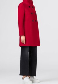 Cappotto rosso in lana con bottoni neri doppi e tasca, dotato di colletto alto. Abbinato a pantaloni neri a gamba larga e sneakers bianche.
