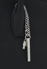 Borsa a tracolla nera in materiale sintetico con superficie texture, dotata di catena e portachiavi in argento con un'etichetta "Lacoste".
