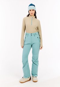 Beige lange mouw top met ritskraag; aqua skibroek met voorzakken en knoopsluiting; crème sneeuwlaarzen; blauwe gebreide beanie met donkere accenten.