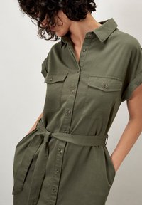 Olivgrünes, kurzärmeliges Hemdkleid mit Knopfleiste, zwei Brusttaschen, Gürtel in der Taille und einer glatten, weichen Textur.
