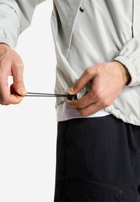Χέρια που ρυθμίζουν το κορδόνι ενός ανοιχτόχρωμου γκρι μπουφάν που φοριέται πάνω από άσπρο πουκάμισο και μαύρο παντελόνι.