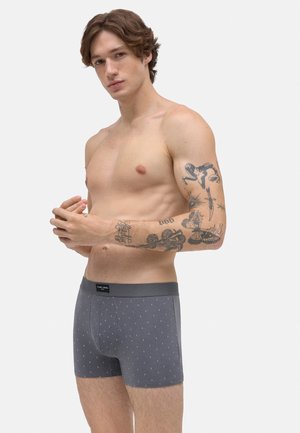 Hombre sin camisa con tatuajes en el brazo derecho vistiendo bóxer gris con estampado, de pie contra un fondo blanco liso.