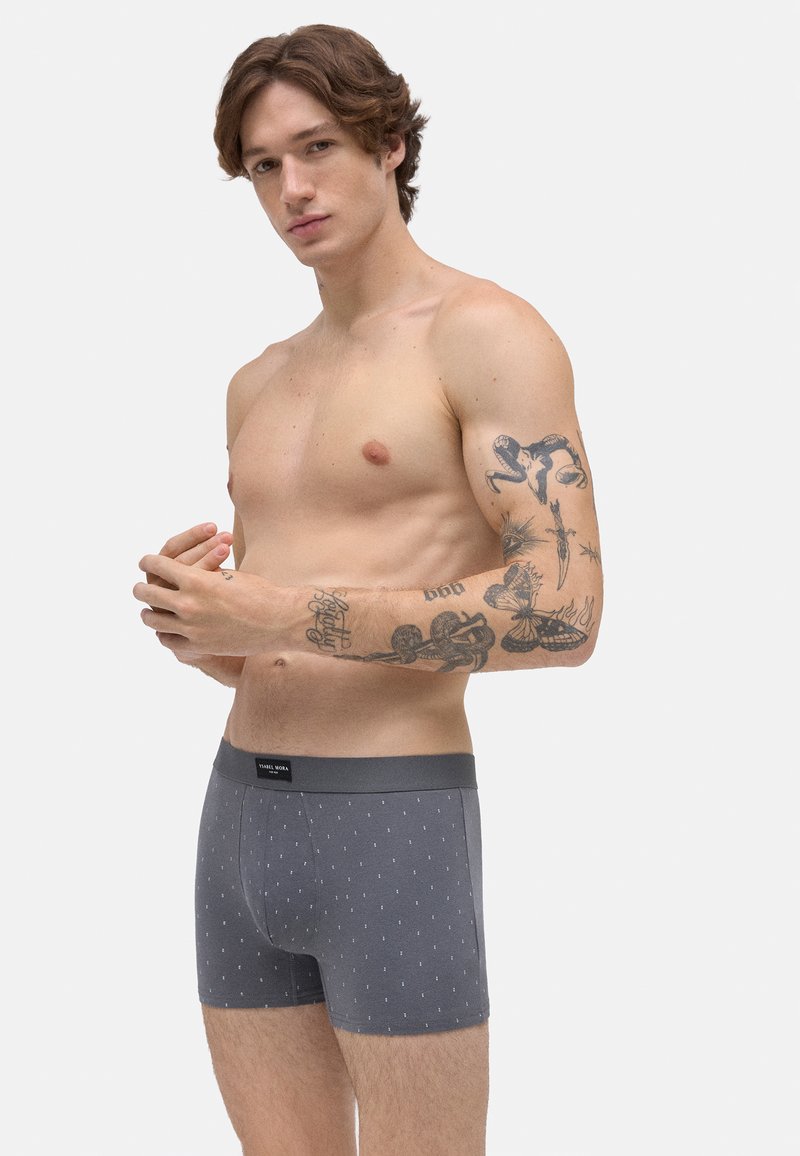 Calzoncillos tipo bóxer grises con un patrón de puntos, hechos de tela suave. Presentan una cintura cómoda y un diseño ajustado que resalta contornos definidos.