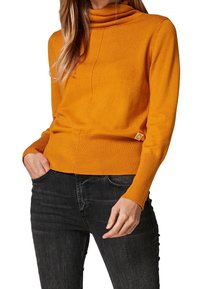 Orange turtleneck sweater made of soft knit material with a fitted design, ribbed hem, and side logo detail. Paired with dark jeans.