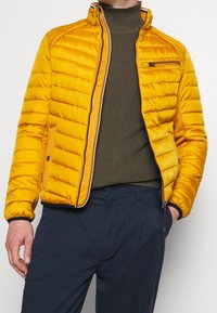 Gul puffjacka med vertikal quiltad design, dragkedja fram och sidofickor. Har en hög krage och mörka kontrasterande detaljer.