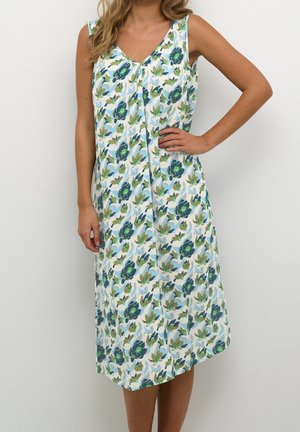 Femme portant une robe midi blanche sans manches à motifs de fleurs bleues et vertes, debout avec une main sur la hanche.