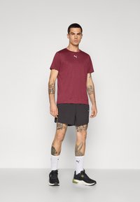 Burgunderfarbenes Sport-T-Shirt mit kurzen Ärmeln und weißem Puma-Logo, kombiniert mit schwarzen Shorts, weißen Socken und schwarzen Laufschuhen.