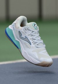 Scarpa sportiva bianca con tomaia texturizzata, dettagli blu e suola turchese. Presenta un design con lacci e un marchio distintivo sulla linguetta.