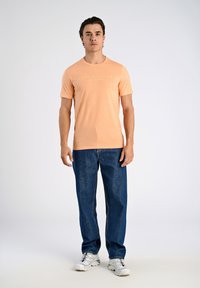 Camiseta de manga corta color durazno con el texto "COPENHAGEN LINDENBERGH", combinada con jeans sueltos de color azul oscuro y zapatillas blancas. Diseño simple, corte casual.