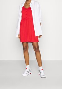 Robe rouge sans manches avec un drapé à la taille, fermeture par bouton, associée à une chemise blanche et des baskets grises et blanches avec des accents rouges.