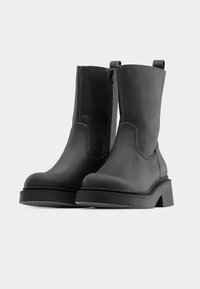 Ζευγάρι μαύρες δερμάτινες μπότες μέχρι τη μέση της γάμπας με χοντρές σόλες και πλαϊνά φερμουάρ, εμφανίζονται σε απλό λευκό φόντο.