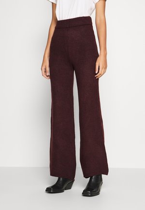 Vrouw die donkerbruine broek met hoge taille en wijd uitlopende pijpen draagt, gemaakt van een textuurgebreide stof, gecombineerd met zwarte enkellaarsjes met hak.