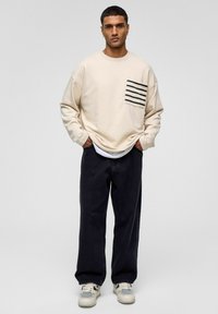 Beige sweatshirt met een gestreepte zak, gecombineerd met donkere jeans en lichte sneakers. De sweatshirt heeft een losse pasvorm en lange mouwen.