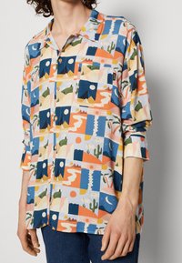 Chemise à manches courtes à boutons en tissu léger présentant un motif géométrique coloré avec des montagnes, des cactus et des motifs de soleil.