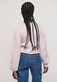 Vaaleanpunainen, teksturoitu neulepusero, jossa on ilmavat hihat, yhdistetty korkeavyötäröisiin sinisiin farkkuihin. Näkymä takaa, esitellen pitkät letit.