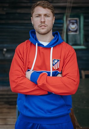 Jonge man met kort haar die een rood-blauwe hoodie met het logo van Atlético Madrid draagt, staand met gekruiste armen binnen.