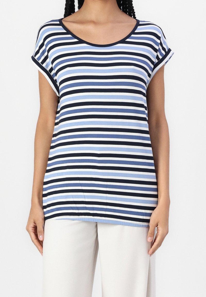 Femme portant une chemise ample à manches courtes avec des rayures horizontales bleu marine, bleu clair, noir et blanc, associée à un pantalon blanc.