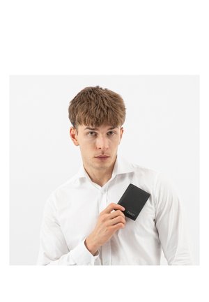 Homme en chemise blanche tenant un portefeuille noir près de sa poitrine sur un fond blanc uni.